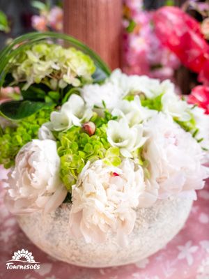 Bouquet de pivoines blanches et hortensias verts dans un vase rond en verre, fond fleuri.